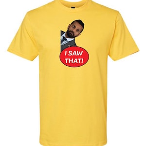 Könnte beinhalten: Ein gelbes T-Shirt mit einem Bild eines Mannes, der um eine Ecke lugt. Der Text "I saw that!" ist in Rot auf einem weißen Kreis gedruckt.