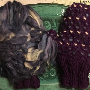 Puede incluir: Una manopla de punto morado con un patrón de corazones blancos y una bola de lana en tonos azules, blancos y morados.