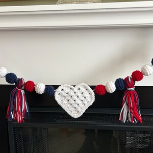 May include: A red, white, and blue crocheted heart garland with white pom poms and red, white, and blue tassels hanging over a fireplace.
