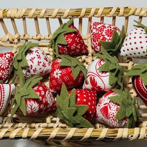 May include: A woven basket filled with fabric strawberries. The strawberries are red and white with various patterns, including polka dots and floral designs. Each strawberry has a green felt leaf and stem.