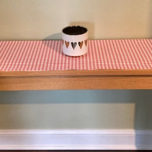 May include: A wooden table with a red and white checkered table runner. A white ceramic container with gold hearts is on the table runner.