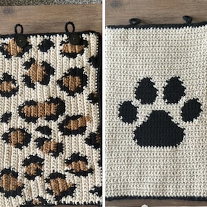 May include: Two rectangular crochet hanging towels featuring loops at the top with black buttons. One towel has a cream, tan, and black leopard print pattern, while the other is cream-colored with a large black paw print design in the center.