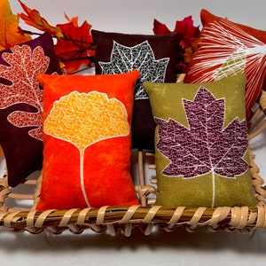 May include: Five decorative throw pillows with different leaf designs. The pillows are in a wicker basket. The leaves are in shades of orange, green, brown, and purple.