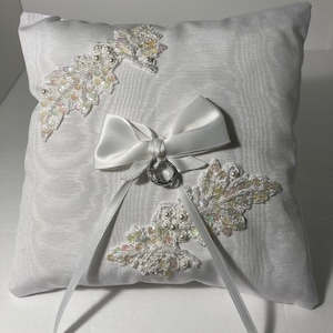 May include: White satin ring pillow with a white satin bow and two pearl and iridescent lace embellishments. A silver ring is placed on the bow.