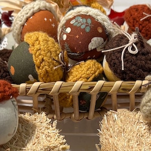 May include: A wicker basket filled with decorative acorn ornaments. The acorns have fabric bodies with various patterns and colors, including orange, brown, and green. Each acorn has a crocheted cap in shades of brown, tan, and orange. The basket sits on a bed of straw.