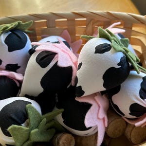 May include: A basket filled with handmade cow print strawberries. The strawberries are made of fabric and have pink felt leaves.