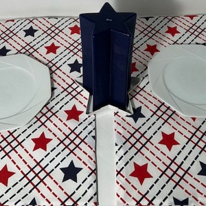 May include: Two white dinner plates with a scalloped edge are placed on a white tablecloth with a red, white, and blue star pattern. A blue star-shaped vase is in the center of the table.