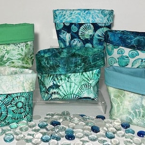 May include: Five fabric storage bins with a sea life theme. The bins are in shades of blue and green with a variety of patterns. The bins are sitting on a white surface with clear glass stones.