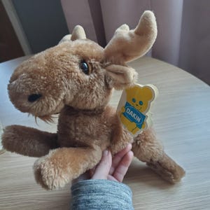 Könnte beinhalten: Ein brauner Plüsch-Elch mit großen Ohren und einem Dakin-Etikett, das an seinem Rücken befestigt ist. Das Etikett ist gelb mit einem blauen Rand und zeigt einen Comic-Bären mit dem Wort "DAKIN" darauf.