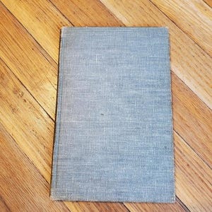 May include: A rectangular book with a light blue cover with a textured, woven appearance. The book is lying on a wooden surface with a diagonal pattern of planks.