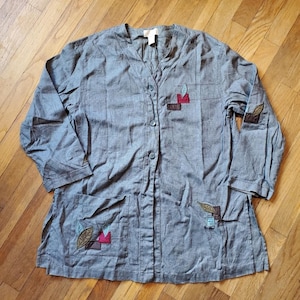 May include: Gray button-down shirt with three-quarter sleeves and two front pockets. The shirt features decorative leaf and geometric patches in shades of brown, red, and green. The shirt is made of a textured fabric.