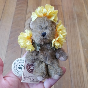 May include: A small, brown teddy bear wearing a crown of yellow flowers. The bear has a black nose and small, round ears. A tag with the text "BE CALM" and "BOYDS" is attached. The bear is a collectible item.