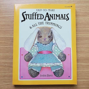 May include: A book titled "Easy-to-Make Stuffed Animals & All the Trimmings" features a plush bunny wearing a blue dress. The cover is yellow with a pink and white border. The author's name, Jodie Davis, is printed at the bottom.