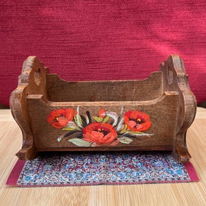 Könnte beinhalten: Eine kleine Wiege aus Holz mit geschwungenen Kufen und herzförmigen Ausschnitten an den Seitenteilen. Die Vorderseite ist mit einem handgemalten Blumenmotiv aus leuchtend roten Mohnblumen und grünen Blättern verziert und steht auf einem Teppich.