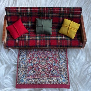 Könnte beinhalten: Miniatur-Puppenhaus-Möbelset mit einem rot karierten Sofa mit roten, grünen und gelben Kissen. Ein kleiner gemusterter Teppich in Rot, Blau und Weiß ist vor dem Sofa platziert. Das Set ist für ein Puppenhaus.