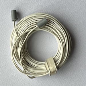 May include: Coiled cream-coloured electrical wire with two grey connectors and a beige clip. The wire is arranged in a circular pattern. The connectors are cylindrical, and the clip secures the wire, suggesting it's new and ready for use.