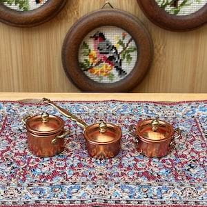 May include: Three miniature copper-colored pots with lids and gold-toned handles, arranged on a rectangular rug featuring a detailed red, blue, and white floral pattern. The pots have a metallic finish and small, rounded side handles.