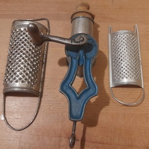May include: A vintage kitchen grater set featuring a central hand-crank grater with a blue frame and two interchangeable grating plates. The set includes a wooden handle and a metal clamp for securing it to a surface. The graters are made of metal.