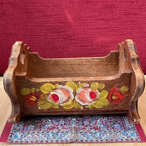 Könnte beinhalten: Eine kleine, rechteckige Wiege aus Holz mit geschwungenen, ausgeschnittenen Seitenteilen. Die Vorderseite ist mit einem handgemalten Blumenmuster aus rosa, weißen und roten Rosen verziert, die von grünen Blättern umgeben sind. Das Holz ist braun.