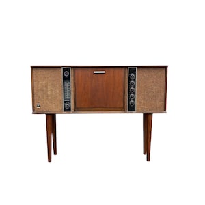 May include: A wooden console stereo cabinet featuring a central compartment with a silver handle, flanked by two textured brown speaker grilles. The unit includes a vertical radio tuner and control knobs on black panels, supported by four tapered legs.