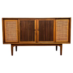 May include: Mid-century modern walnut stereo cabinet with two doors and two side panels with woven cane inserts. The cabinet has a dark wood finish and sits on four tapered legs.
