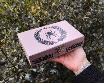 May include: A light pink wooden box with a black spider and crescent moon design inside a wreath. The box has a metal clasp and black leaf designs on the edges. The box is held in a hand.