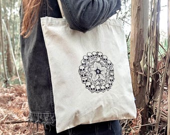 Op de afbeelding: Een crèmekleurige draagtas met een zwart-wit bloemenontwerp omringd door schedels. De tas is gemaakt van een stevige stof en heeft een eenvoudig ontwerp, ideaal voor het dragen van boodschappen of boeken.