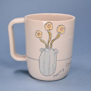 May include: Cream-colored ceramic mug with a large handle. The mug features a hand-drawn illustration of yellow flowers in a blue vase, with the words "Pretty Flowers" written on the side.