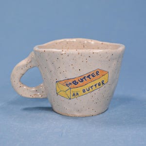 May include: A speckled ceramic mug with a handle. The mug features a hand-drawn illustration of a butter stick with the text "AA BUTTER" in blue and orange. The mug is set against a blue background.