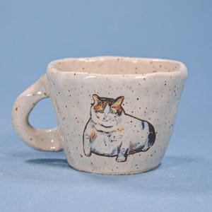 May include: A speckled ceramic mug with a rounded handle. The mug features a hand-painted illustration of a cat with orange, brown, and white fur. The mug has a square-like shape and is set against a light blue background.