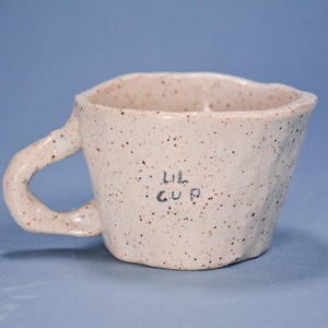 May include: A speckled, off-white ceramic mug with a unique, irregular shape and a curved handle. The mug has the words "LIL CUP" written on the side. The mug is set against a solid blue background.