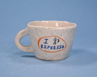May include: A small, speckled ceramic espresso cup with a curved handle. The cup features an orange oval with the words "I Espresso" in blue, along with a small illustration of a coffee maker. The cup is off-white with black speckles.