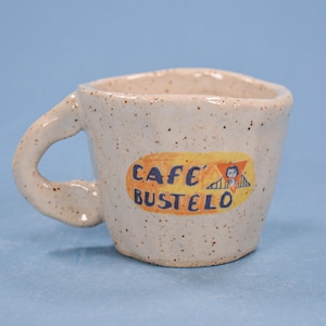 May include: A speckled ceramic coffee mug with a curved handle. The mug features a yellow and orange label with the text "CAFE BUSTELO" and a small illustration. The mug is off-white with dark speckles.