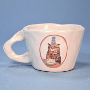 Może przedstawiać: Biały ceramiczny kubek z teksturowanym uchwytem. Kubek posiada kolorową ilustrację kota w czapce urodzinowej i trzymającego ciasto. Ilustracja kota znajduje się w czerwonym okręgu.