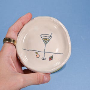 Peut inclure: Une petite coupelle en céramique blanc cassé avec un dessin d'un verre à cocktail, une pêche et une fraise. La coupelle est tenue sur un fond bleu. Le bord de la coupelle est légèrement irrégulier.
