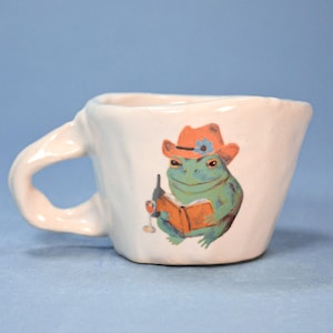 May include: White ceramic mug featuring a frog in a cowboy hat, reading a book and holding a wine glass. The mug has a textured handle and an irregular shape. The frog illustration is in shades of green, orange, and blue, adding a touch of whimsy.