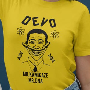 Peut inclure: T-shirt jaune avec le mot "DEVO" et un dessin noir et blanc d'un visage souriant. Le texte "MR.KAMIKAZE MR.DNA" est en dessous. Le design comprend des symboles atomiques et des brins d'ADN.