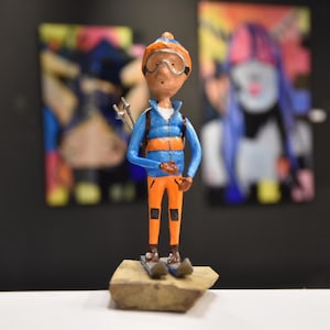 May include: A small figurine of a skier, wearing an orange and blue outfit, goggles, and skis. The figure is standing on a small, rectangular base. The background features two abstract paintings.