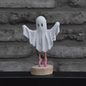 Könnte beinhalten: Eine skurrile weiße Geisterfigur mit ausgestreckten Armen und zwei schwarzen Punktaugen. Der Geist trägt rosa Ballettschuhe und steht auf einem Holzsockel. Der Hintergrund ist eine graue Backsteinmauer.