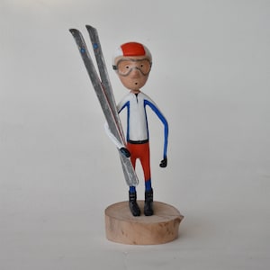 May include: A wooden figurine of a skier wearing a red and white hat, blue and white ski suit, and goggles. The skier is holding two silver skis.