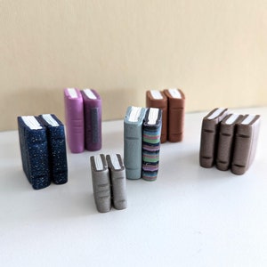 Op de afbeelding: Een verzameling miniatuurboeken gemaakt van polymer clay. De boeken zijn in paren gestapeld en hebben verschillende kleuren en patronen, waaronder blauw, bruin, paars en groen.