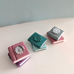 Op de afbeelding: Drie stapels miniatuurboeken met decoratieve omslagen. Het bovenste boek in elke stapel heeft een ander ontwerp: een bloemenpatroon, een bladontwerp en een sneeuwvlokontwerp. De boeken zijn gemaakt van polymer clay en zijn geschilderd in verschillende tinten roze, paars, turquoise en grijs.