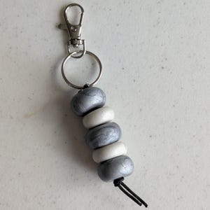 Può includere: Un portachiavi con un moschettone in metallo color argento e un anello circolare collegato a cinque perline alternate. Le perline consistono in tre perline grigio argento metallizzato e due perline bianche unite, infilate su un cordino nero.
