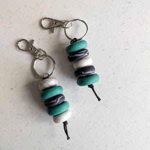 Può includere: Due portachiavi con moschettoni e anelli in metallo argentato. Ogni portachiavi presenta una fila verticale di quattro perline arrotondate in colori alternati: verde acqua, nero con vortici bianchi e bianco solido, rifinito con un cordino nero.
