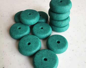 May include: A collection of teal-colored, donut-shaped beads with a speckled texture and a central hole. Some beads are stacked, while others are scattered, all on a speckled white surface. These beads are suitable for jewelry making and crafts.