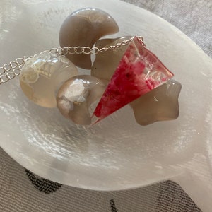 K&ouml;nnte beinhalten: Eine silberne Kettenkette mit einem dreieckigen Anh&auml;nger in Rosa und Wei&szlig;. Der Anh&auml;nger ist aus Harz und hat ein florales Design. Die Kette wird auf einer wei&szlig;en Selenitplatte mit anderen wei&szlig;en Steinen pr&auml;sentiert.