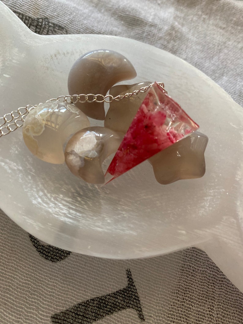 K&ouml;nnte beinhalten: Eine silberne Kettenkette mit einem dreieckigen Anh&auml;nger in Rosa und Wei&szlig;. Der Anh&auml;nger ist aus Harz und hat ein florales Design. Die Kette wird auf einer wei&szlig;en Selenitplatte mit anderen wei&szlig;en Steinen pr&auml;sentiert.