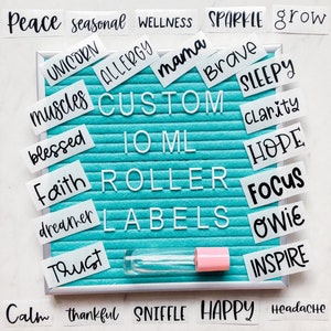 May include: A turquoise letter board with white letters spelling out "CUSTOM 10 ML ROLLER LABELS".  The board is surrounded by white labels with black text, including words like "peace", "unicorn", "mama", "brave", "sleepy", "clarity", "hope", "focus", "owie", "inspire", "calm", "thankful", "sniffle", and "happy".