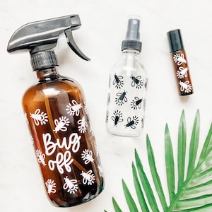 May include: Three glass bottles with black and white bee designs. The bottles are labeled "Bug Off" and are filled with a clear liquid. The bottles are on a white surface with a green leaf in the background.