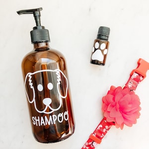 May include: A brown glass bottle with a pump dispenser and a white dog silhouette with the word "Shampoo" printed on the bottle. A small brown glass bottle with a white paw print and a red and white dog collar with a red flower are also in the image.